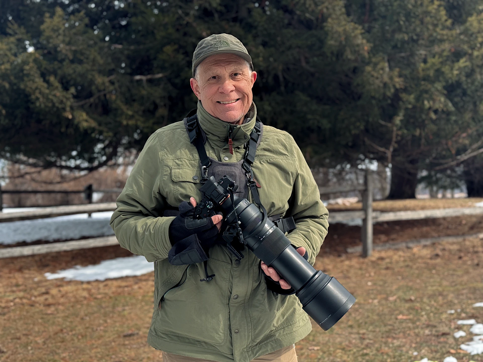 A man in an olive green jacket and cap holds a professional camera with a large telephoto lens outdoors near trees and patchy snow.