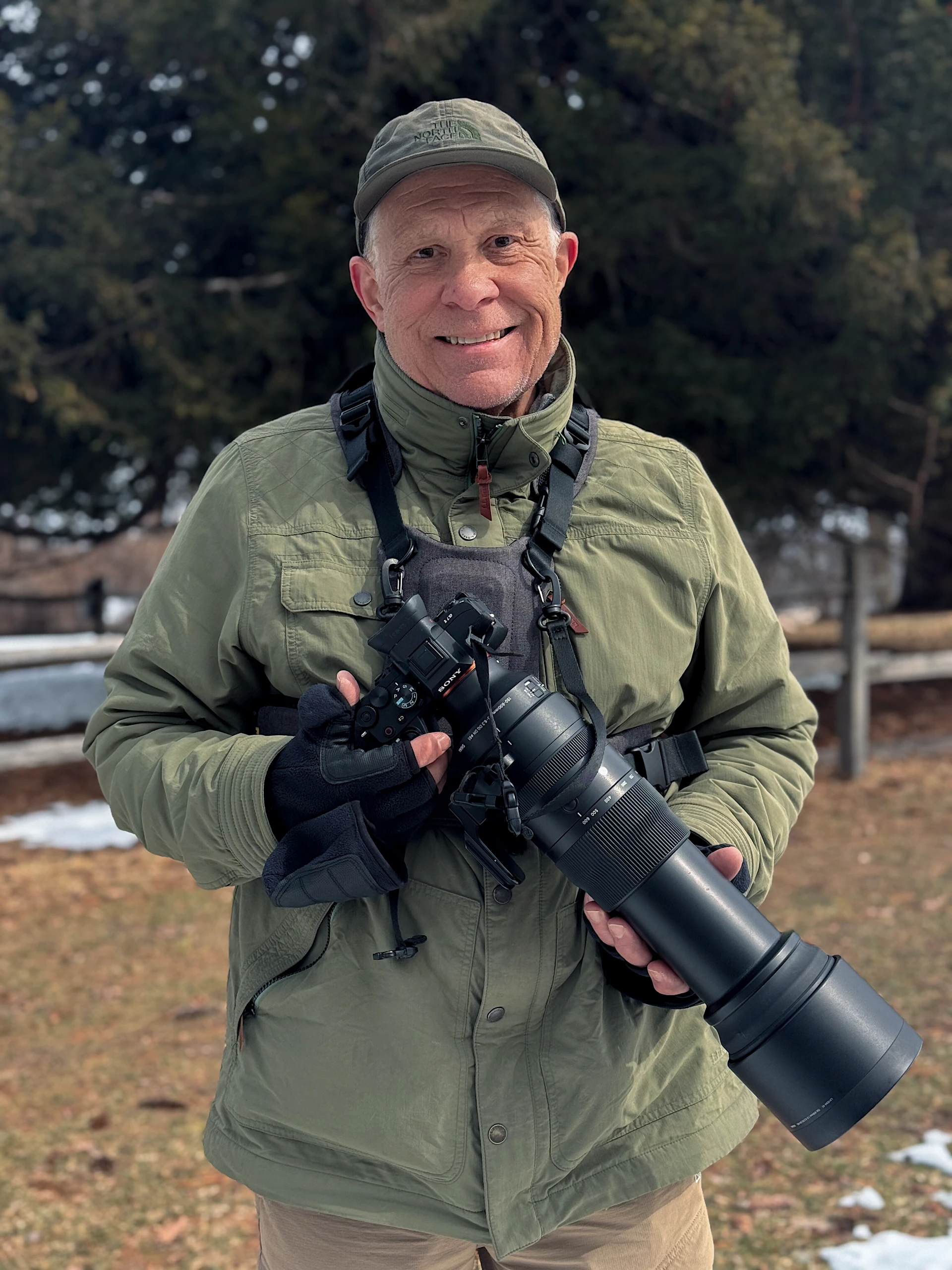 A man in a green jacket and North Face cap holds a Sony camera with a large telephoto lens outdoors in a cold, partially snow-covered setting. A man in a green jacket and North Face cap holds a Sony camera with a large telephoto lens outdoors in a cold, partially snow-covered setting.